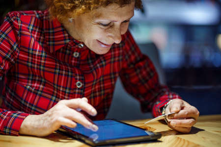 Senior woman ordering Christmas gifts online on digital tablet in coffee shop. Mature buyer holding tablet pc and credit card. Elderly woman shopping online. Online personal finance. Black Friday.の写真素材