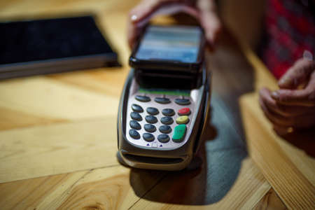 Mature female doing POS terminal and smartphone payment transaction in evening at coffee shop table. Senior customer using smartphone to pay for her cafe purchase. Woman making payment in cafe.の写真素材
