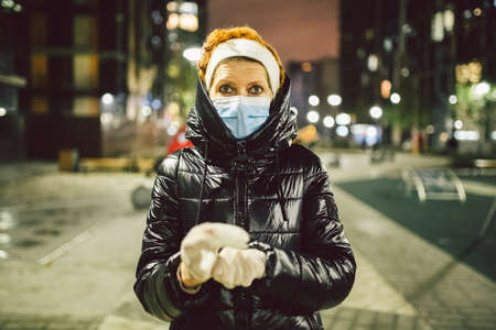 Senior Caucasian red hair woman medical protective mask on her face and white gloves during the coronavirus pandemic quarantine on street European city looks at camera, portrait background cityview.の写真素材