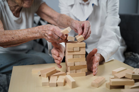 Mature doctor conducting session, therapy for senior patient in nursing home, training fine motor skills for dementia, alzheimer disease and recovery institute by folding wooden blocksの写真素材