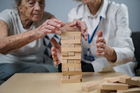Dementia therapy in playful way, training fingers and fine motor skills, build wooden blocks into tower. Senior woman 90 years old and doctor playing educational game in nursing home.の写真素材