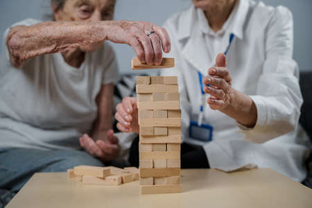 Mature doctor conducting session, therapy for senior patient in nursing home, training fine motor skills for dementia, alzheimer disease and recovery institute by folding wooden blocksの写真素材