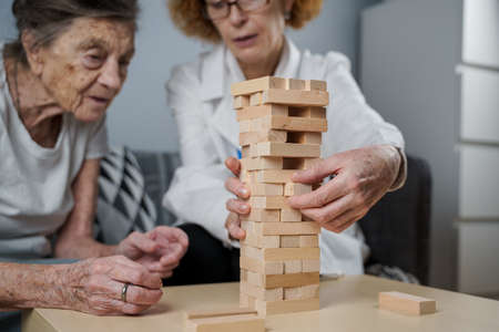 Dementia therapy in playful way, training fingers and fine motor skills, build wooden blocks into tower, playing . Senior woman 90 years old and doctor playing educational game in nursing home.の写真素材