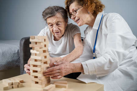 Theme is dementia, aging and games for old people. Caucasian senior woman builds tower of wooden blocks with the help of a doctor as part of a therapy and game at a patient's home.の写真素材