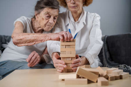 Theme is dementia, aging and games for old people. Caucasian senior woman builds tower of wooden blocks with the help of a doctor as part of a therapy and game at a patient's home.の写真素材