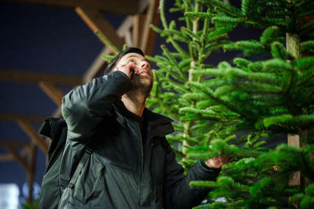 Father buys Christmas tree and talks on phone. Purchase of timber at market. Bazaar. Christmas sale. Satisfied with the choice of spruce. Christmas shopping and preparation for the celebration.の写真素材