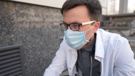 Caucasian young doctor man sits down on the stairs near the clinic building, tired and unhappy rubbing his nose and eyes, feeling tired and headache. Health care worker stress and frustration concept.の写真素材