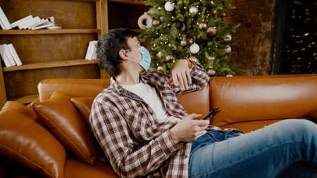 Sad man in mask surfing Internet alone at home on couch at Christmas time. Quarantine during New Year holidays ruined mood and introduced into depression, communication with friends and family online.の写真素材