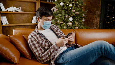 Sad man in mask surfing Internet alone at home on couch at Christmas time. Quarantine during New Year holidays ruined mood and introduced into depression, communication with friends and family online.の写真素材