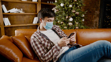 Young sad man in medical mask reads news from smartphone at home on couch near Christmas tree in quarantine due to virus. Bad news, illness.の写真素材