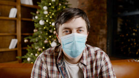 Unpleasant mood for Christmas holidays. Sad unhappy handsome man sits on the sofa wearing a mask. Feeling of hopelessness lockdown, quarantine virus.の写真素材