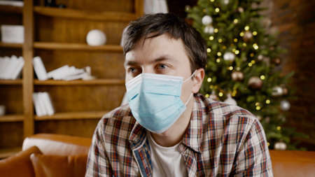 Upset with bad mood man in protective mask is suffering from loneliness at home while sitting on couch near Christmas tree during New Year holidays.の写真素材