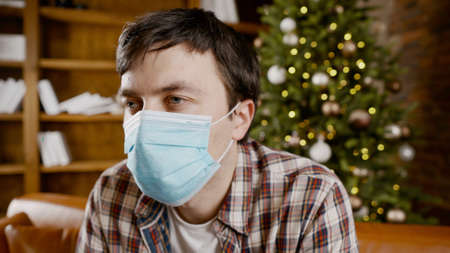 Upset with bad mood man in protective mask is suffering from loneliness at home while sitting on couch near Christmas tree during New Year holidays.の写真素材
