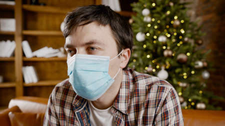 Unpleasant mood for Christmas holidays. Sad unhappy handsome man sits on the sofa wearing a mask.の写真素材