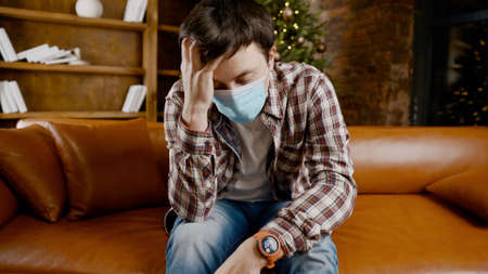 Upset with bad mood man in protective mask is suffering from loneliness at home while sitting on couch near Christmas tree during New Year holidays.の写真素材