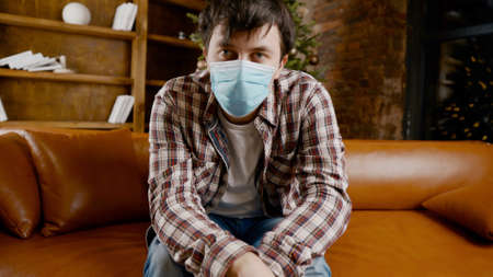 Upset with bad mood man in protective mask is suffering from loneliness at home while sitting on couch near Christmas tree during New Year holidays. New norm, lockdown and quarantine virus.の写真素材