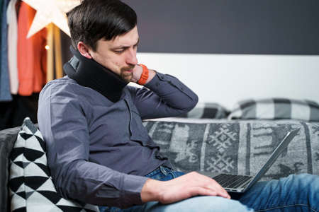 Man employee after accident working at home behind laptop and feels pain in neck, uses black neck collar support. Male with neck injury freelance programmer on sick leave working at computer on couch.の写真素材