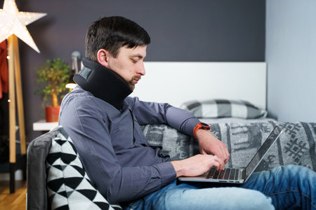 Man employee after accident working at home behind laptop and feels pain in neck, uses black neck collar support. Male with neck injury freelance programmer on sick leave working at computer on couch.の写真素材