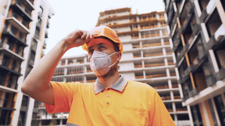 Topic is health protection in production and construction. Caucasian male worker in orange uniform and protective respirator kn 95 puts on construction orange hard hat and poses at construction site.の写真素材