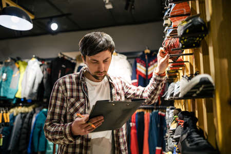 Business owner in sports store works with showcase trekking and mountaineering boots for sale, holding clipboard in his hands, checks availability of goods and collects to process online orders.の写真素材