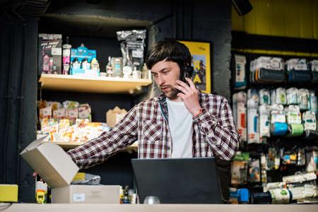 Bike shop owner at work. Store employee takes an order by mobile phone at table near laptop in travel and sporting goods store. Small business, communication, people and service concept.の写真素材