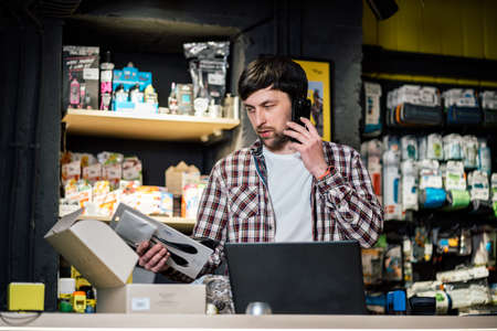 Man takes order by phone in his sporting goods store for sports and tourism sitting at table near computer talking on smartphone. Theme is small business. Sales clerk on phone in bike shop.の写真素材