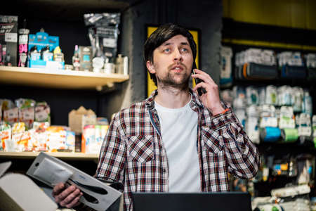 Man takes order by phone in his sporting goods store for sports and tourism sitting at table near computer talking on smartphone. Theme is small business. Sales clerk on phone in bike shop.の写真素材