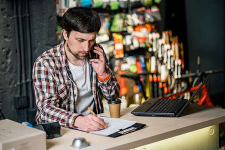 Man takes order by phone in his sporting goods store for sports and tourism sitting at table near computer talking on smartphone. Theme is small business. Sales clerk on phone in bike shop.の写真素材