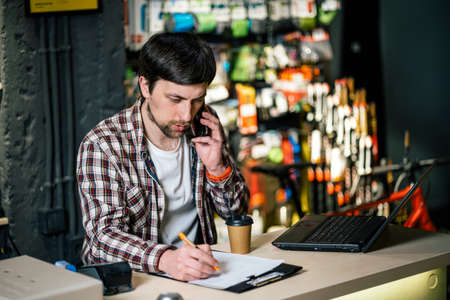 Bike shop owner at work. Store employee takes an order by mobile phone at table near laptop in travel and sporting goods store. Small business, communication, people and service concept.の写真素材