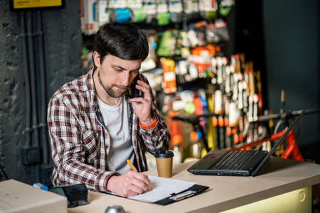 Small business owner on phone and computer in store. Sport shop worker making call with cell phone. Salesman working and talking on cell phone in bicycle shop. Seller taking orders by phone.の写真素材