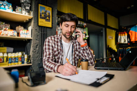 Small business owner on phone and computer in store. Sport shop worker making call with cell phone. Salesman working and talking on cell phone in bicycle shop. Seller taking orders by phone.の写真素材
