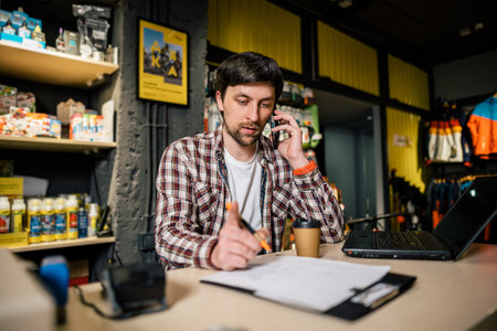 Man takes order by phone in his sporting goods store for sports and tourism sitting at table near computer talking on smartphone. Theme is small business. Sales clerk on phone in bike shop.の写真素材