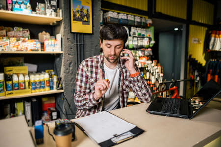 Man takes order by phone in his sporting goods store for sports and tourism sitting at table near computer talking on smartphone. Theme is small business. Sales clerk on phone in bike shop.の写真素材