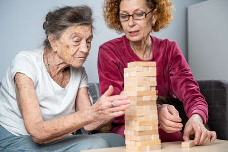 Retired mother and daughter spend time together at home, playing board game and caressing dachshund dog. Caucasian senior woman builds tower of wooden blocks with volunteer assistant in nursing homeの写真素材
