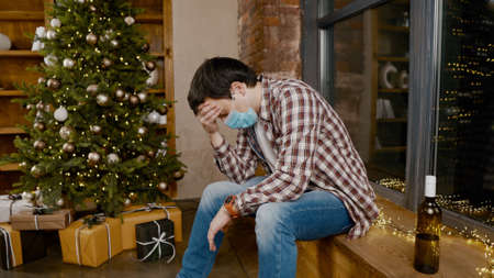 Problems of alcoholism in loneliness, sadness and melancholy during pandemic  Male removes mask from virus, drinking wine from bottle during new year holidays sitting by Christmas treeの写真素材