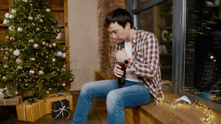 Lonely man wearing  face mask very upset and drunk, sitting by window and Christmas tree and drinking alcohol from bottle. Topic alcohol addiction, depression during quarantine virusの写真素材