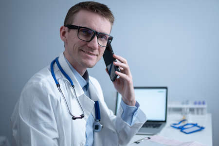 European doctor in white medical coat and glasses consults patient on cell phone in clinic office, sitting at table with laptop. German general practitioner at workplace in hospital talking on phoneの写真素材