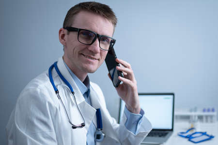 Smiling doctor having phone call at desk. Physicianr talking with his patient in office clinic sitting at table with laptop. German doctor in white lab coat works hospital, consults online by phoneの写真素材