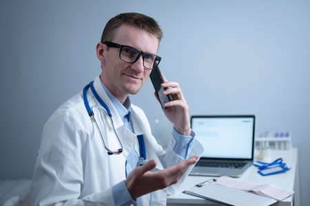 European doctor in white medical coat and glasses consults patient on cell phone in clinic office, sitting at table with laptop. German general practitioner at workplace in hospital talking on phoneの写真素材