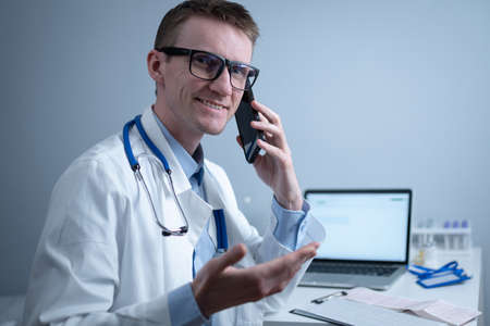 Smiling doctor having phone call at desk. Physicianr talking with his patient in office clinic sitting at table with laptop. German doctor in white lab coat works hospital, consults online by phoneの写真素材