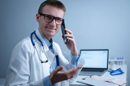 European doctor in white medical coat and glasses consults patient on cell phone in clinic office, sitting at table with laptop. German general practitioner at workplace in hospital talking on phoneの写真素材