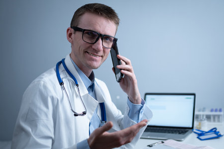 Smiling doctor having phone call at desk. Physicianr talking with his patient in office clinic sitting at table with laptop. German doctor in white lab coat works hospital, consults online by phoneの写真素材