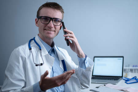 European doctor in white medical coat and glasses consults patient on cell phone in clinic office, sitting at table with laptop. German general practitioner at workplace in hospital talking on phoneの写真素材