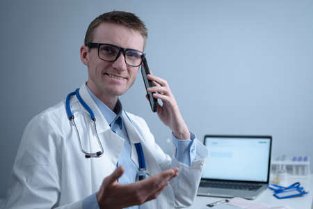 European doctor in white medical coat and glasses consults patient on cell phone in clinic office, sitting at table with laptop. German general practitioner at workplace in hospital talking on phoneの写真素材