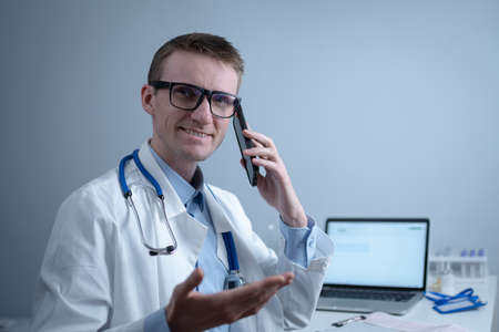 European doctor in white medical coat and glasses consults patient on cell phone in clinic office, sitting at table with laptop. German general practitioner at workplace in hospital talking on phoneの写真素材