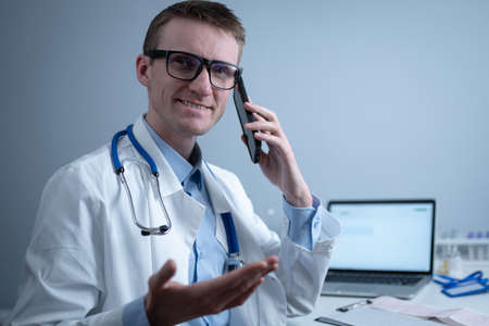 Smiling doctor having phone call at desk. Physicianr talking with his patient in office clinic sitting at table with laptop. German doctor in white lab coat works hospital, consults online by phoneの写真素材