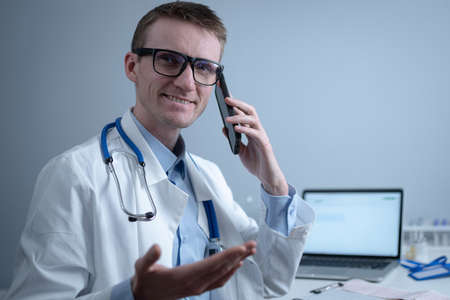 European doctor in white medical coat and glasses consults patient on cell phone in clinic office, sitting at table with laptop. German general practitioner at workplace in hospital talking on phoneの写真素材