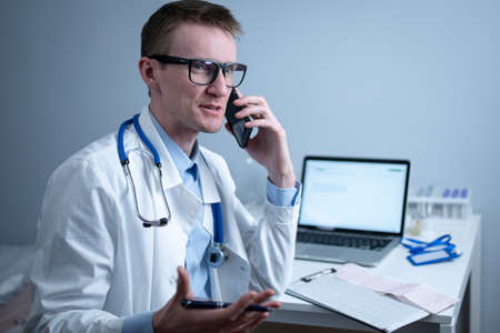European doctor in white medical coat and glasses consults patient on cell phone in clinic office, sitting at table with laptop. German general practitioner at workplace in hospital talking on phoneの写真素材