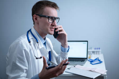 Smiling doctor having phone call at desk. Physicianr talking with his patient in office clinic sitting at table with laptop. German doctor in white lab coat works hospital, consults online by phoneの写真素材