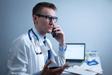 European doctor in white medical coat and glasses consults patient on cell phone in clinic office, sitting at table with laptop. German general practitioner at workplace in hospital talking on phoneの写真素材